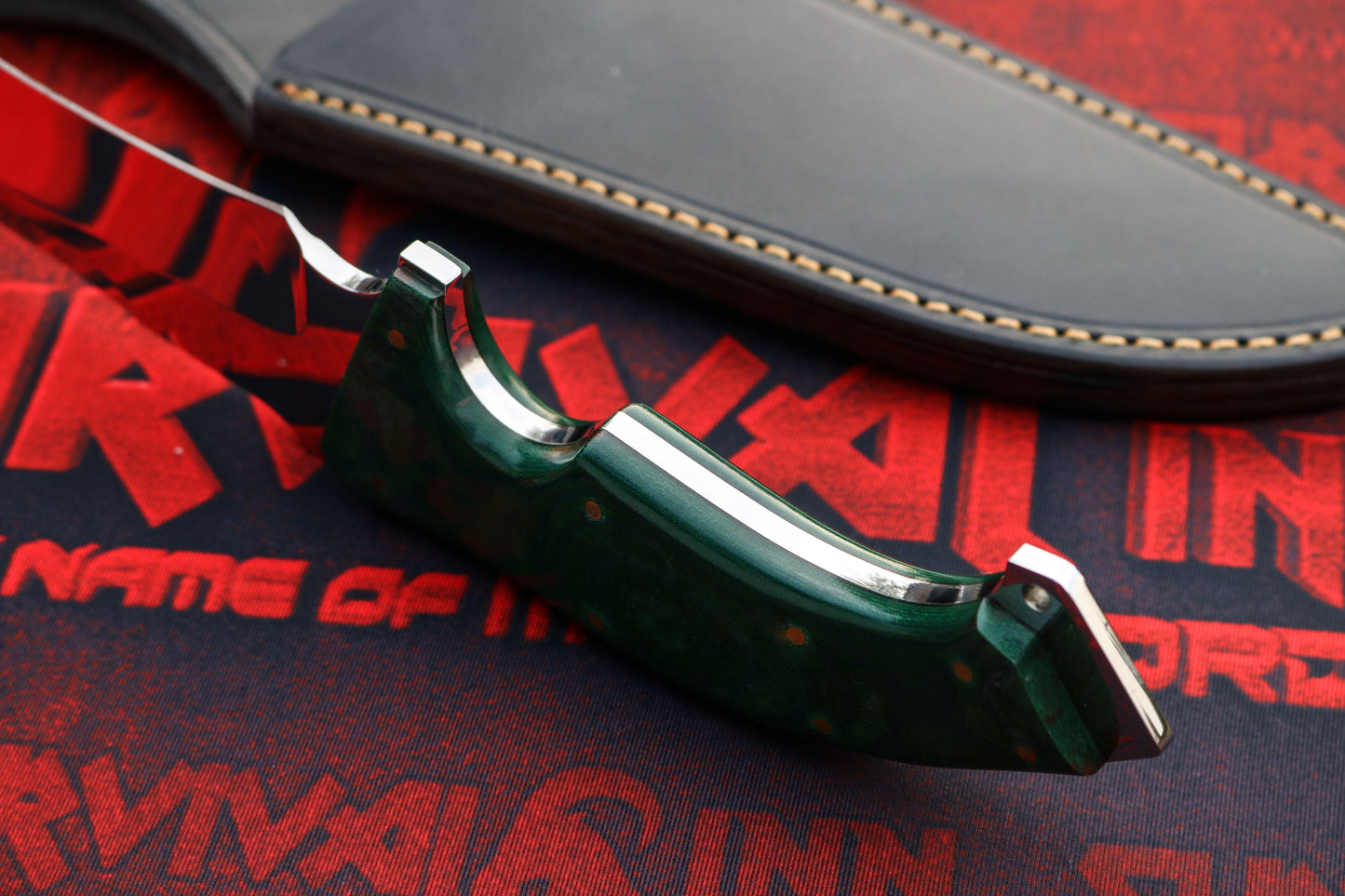 Close-up of handmade tactical Bowie knife handle showing ergonomic green handle scales with finger grooves, metal guard, and polished spine, with leather sheath in background